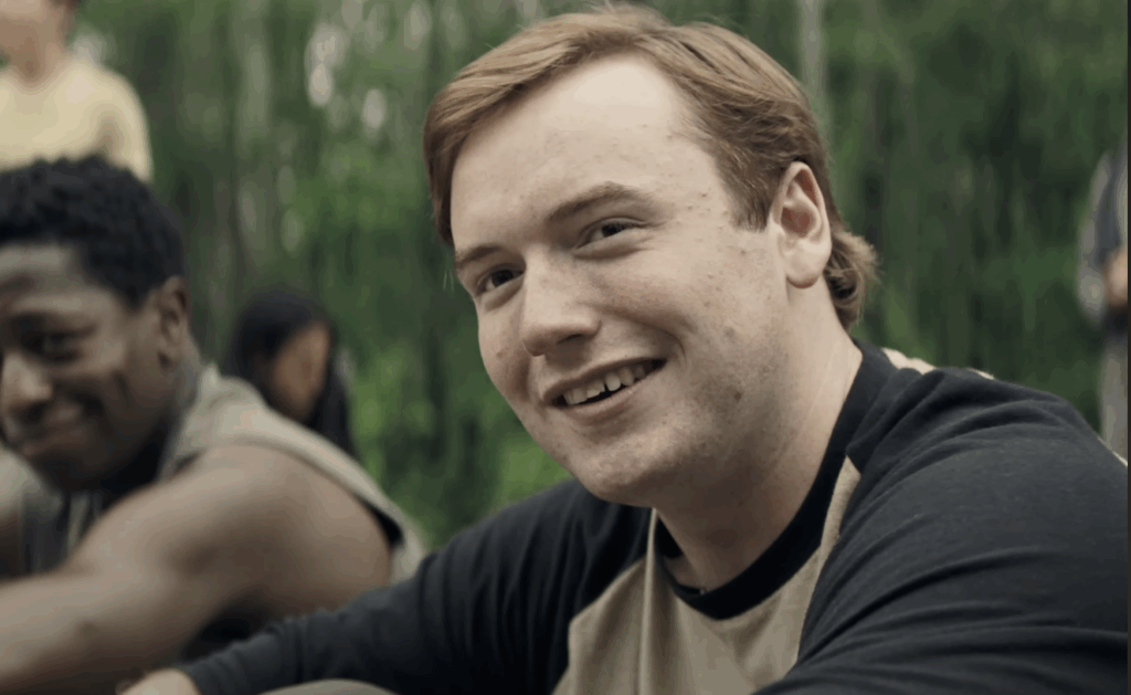 Cooper Hoffman, wearing a black and white T-shirt, and smiling while sitting down on the road in The Long Walk.