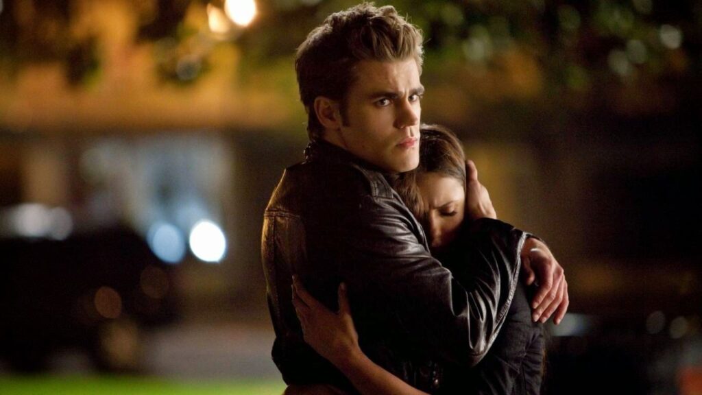 Stefan and Elena embracing outdoors at night, with Stefan holding Elena protectively as warm lights blur in the background.