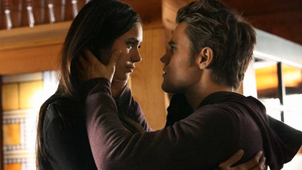 Elena and Stefan in an intimate close-up moment indoors, facing each other with intense eye contact in warm lighting.
