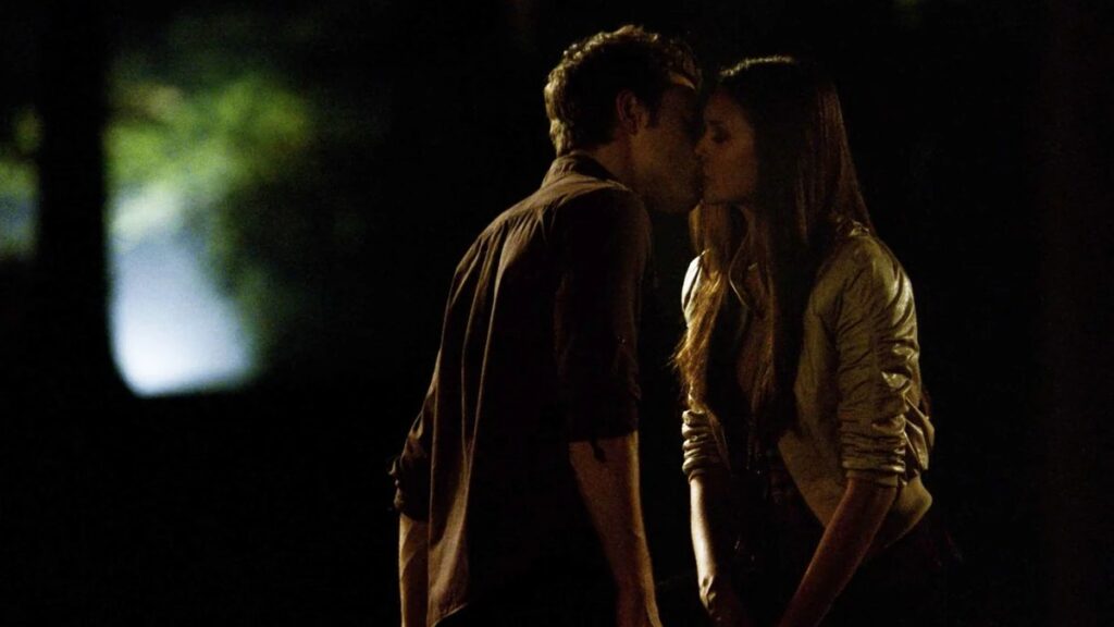 Elena and Stefan sharing a kiss outdoors at night with soft lighting and foliage in the background.