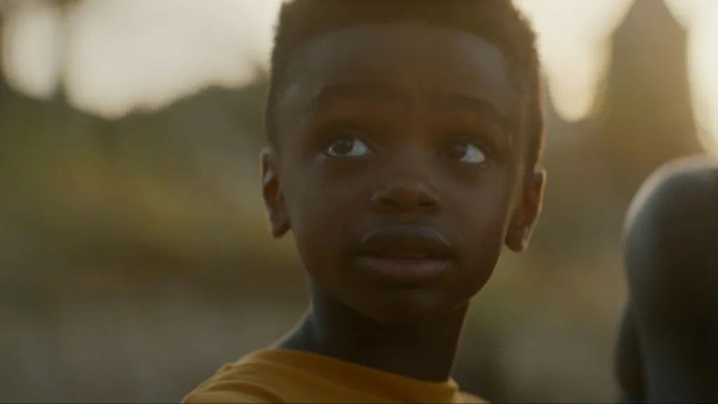 A young Toussaint (T'Challa's son) wearing a bright yellow shirt, looking directly at the camera with a serious expression in an outdoor setting with golden lighting.