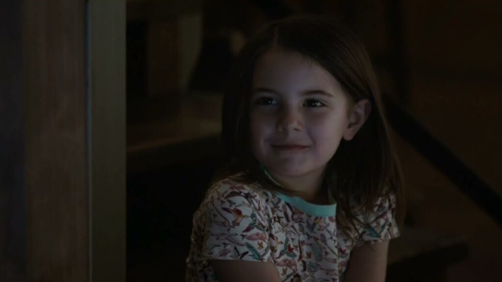 Young Morgan Stark with long hair wearing a patterned shirt, looking towards the camera with a slight smile in a dimly lit indoor setting.