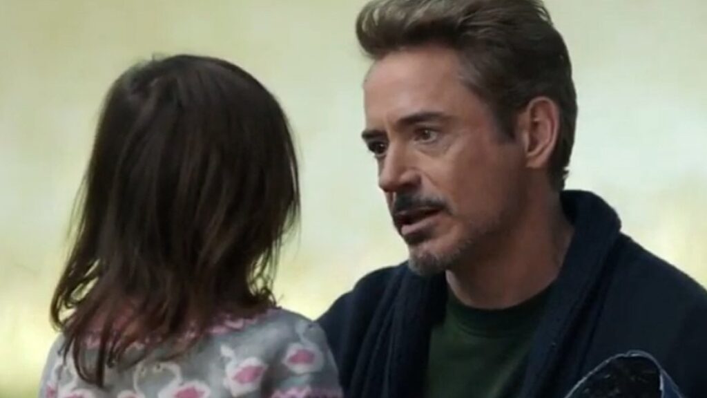 Tony Stark / Iron Man holding his young daughter Morgan Stark, who is wearing a floral patterned outfit, both looking off to the side in a tender father-daughter moment.