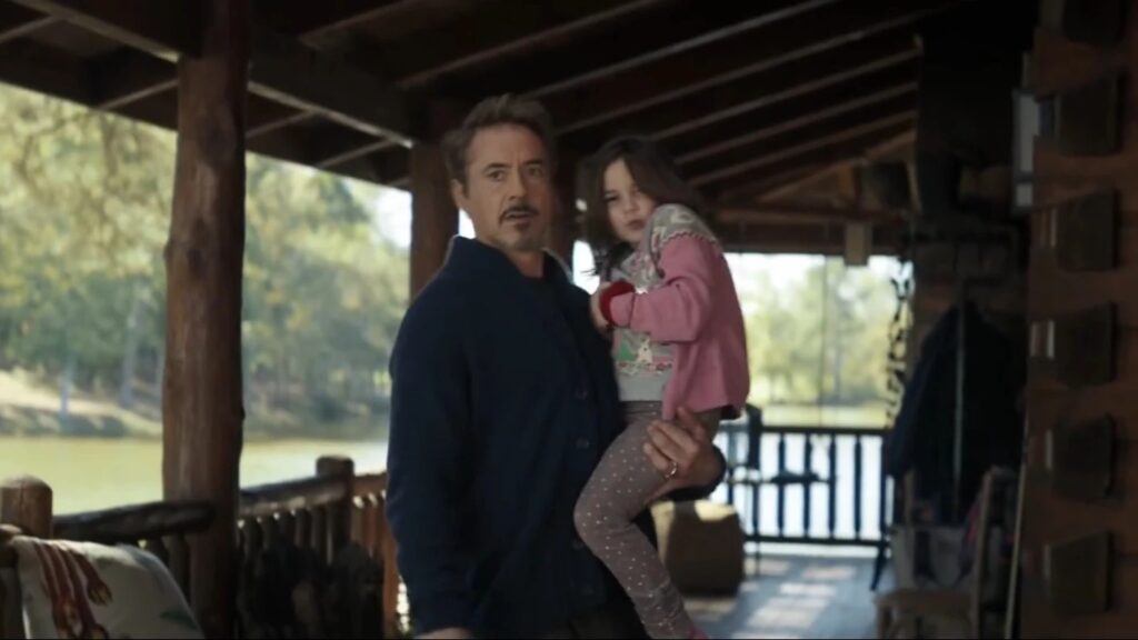 Tony Stark standing on a porch holding young Morgan Stark in his arms, both in casual clothing with Morgan wearing polka-dotted pajamas, in a peaceful family moment.