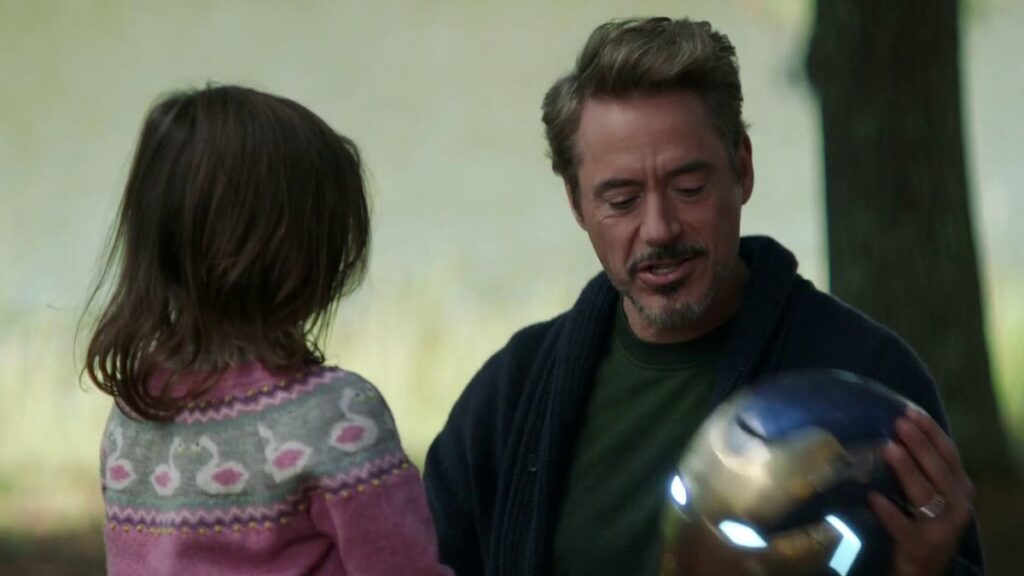 Tony Stark showing young Morgan Stark his Iron Man helmet, which is glowing with blue light, as they sit together in their home.