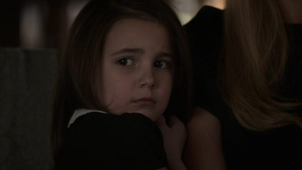 A close-up of young Morgan Stark looking pensively off to the side, wearing dark clothing in a dimly lit setting.