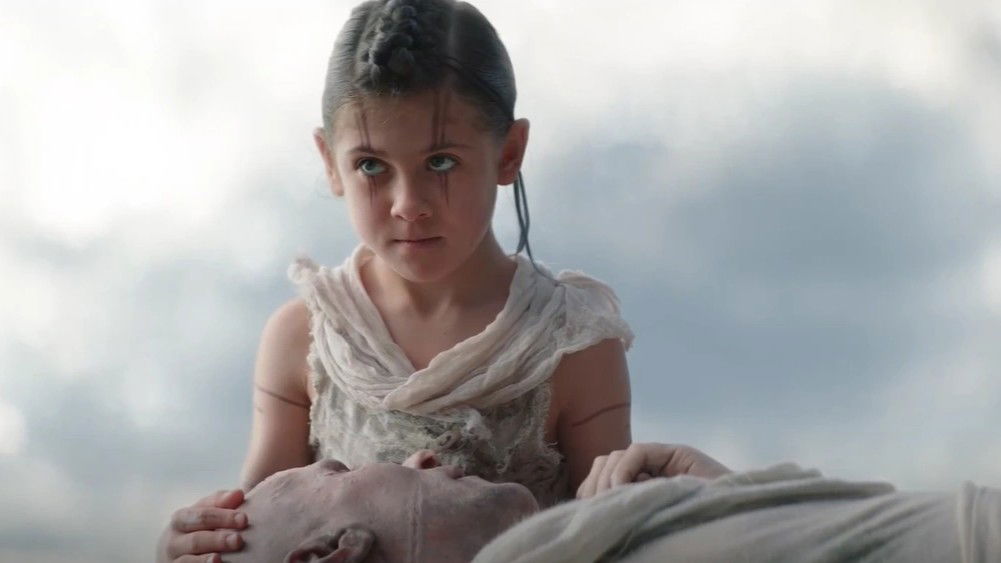 Love as a young girl with dark hair in braids, wearing white/cream colored clothing, looking directly at the camera with an intense expression against a cloudy sky background while Gorr is lying on her lap.
