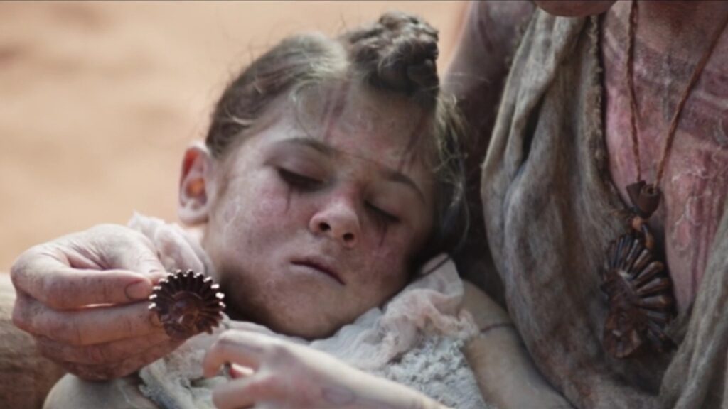 Love lying in Gorr's arms, appearing ill, with intricate face markings, wearing light-colored clothing in an emotional scene from Thor: Love and Thunder.
