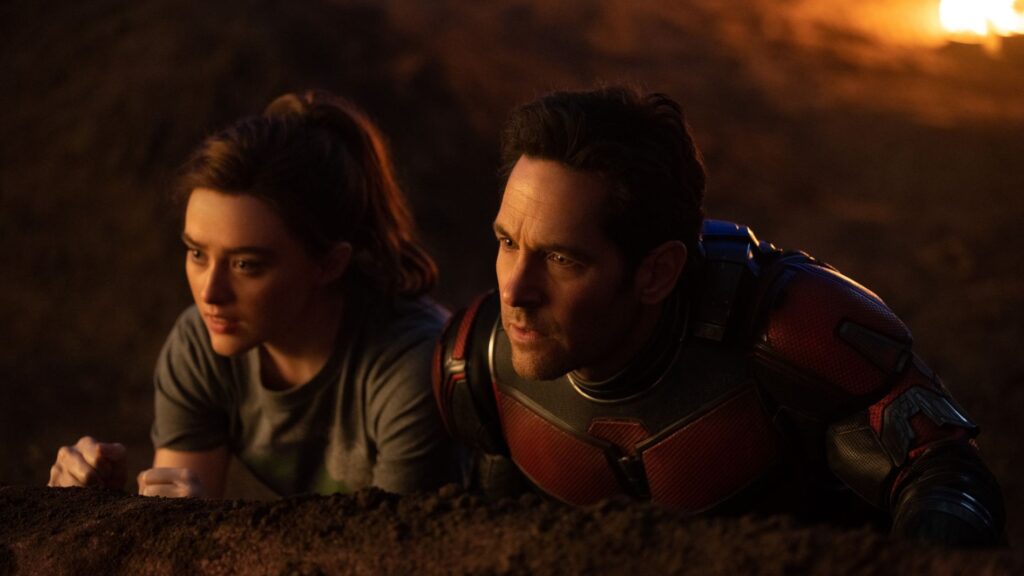 Scott Lang / Ant-Man in his red and black Ant-Man suit lying prone next to his teenage daughter Cassie Lang in casual clothes, both looking ahead intensely in a tense or surveillance situation.