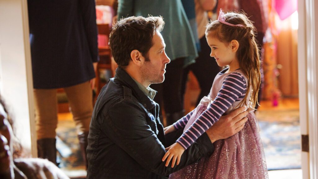 Scott Lang / Ant-Man in casual clothing kneeling down to interact with his young daughter Cassie, who is wearing a pink dress with a striped shirt and small tiara, in a warm, indoor setting.