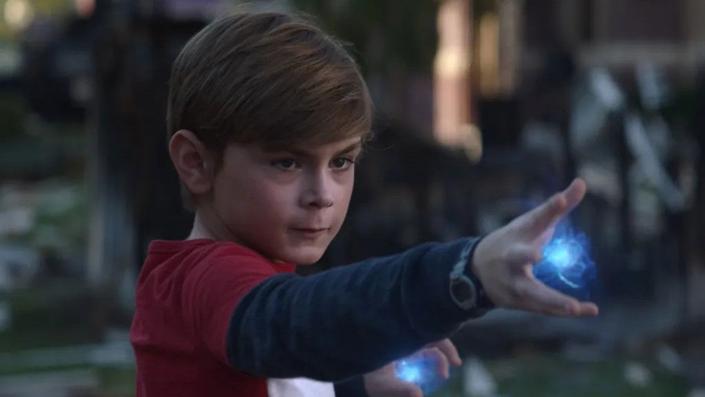 Billy Maximoff from Agatha All Along as a young boy with blonde hair, wearing a red and dark blue outfit, with blue magical energy emanating from his hands as he demonstrates his reality-warping powers.