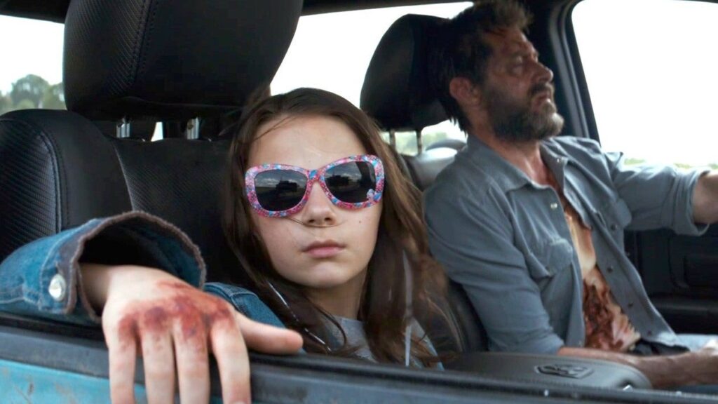 X-23 / Laura Kinney in a car wearing distinctive pink sunglasses, sitting in the passenger seat with Logan visible in the background driving.