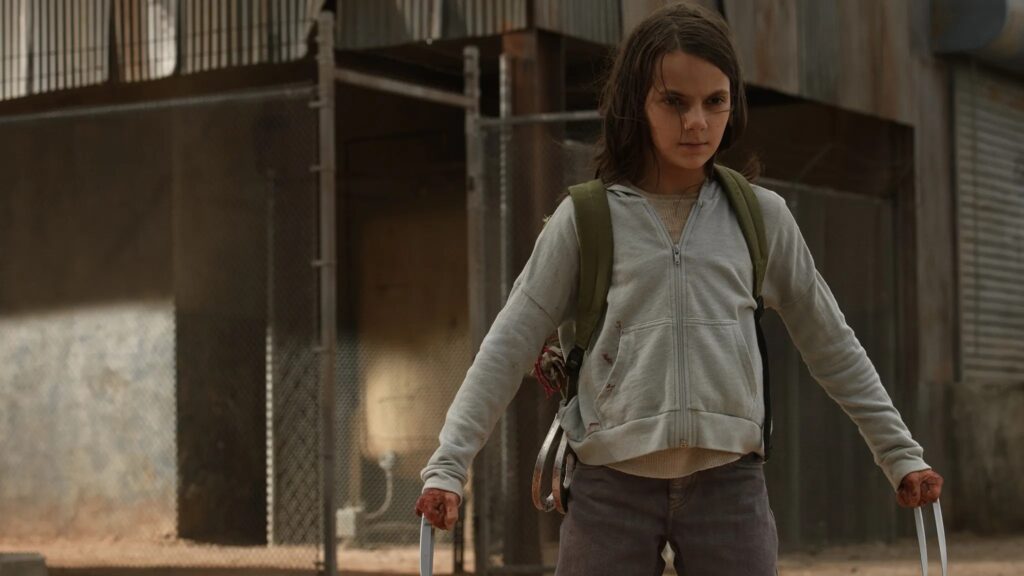 Young X-23 / Laura Kinney as a child in Logan, wearing a gray hoodie and looking serious, with her claws extended from her hands.