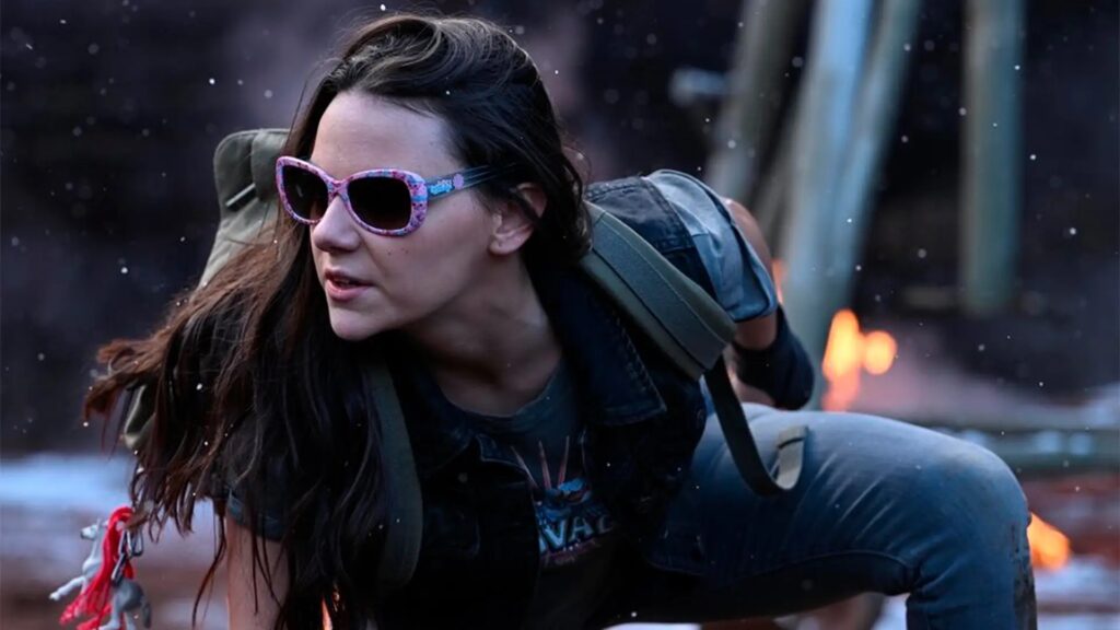 An older X-23 / Laura Kinney in Deadpool & Wolverine, wearing the same pink sunglasses from Logan, in an action scene with explosions in the background.