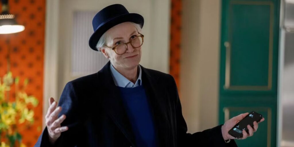 Sazz Pataki in Only Murders In The Building dressed in a black coat, blue sweater, and a hat with glasses