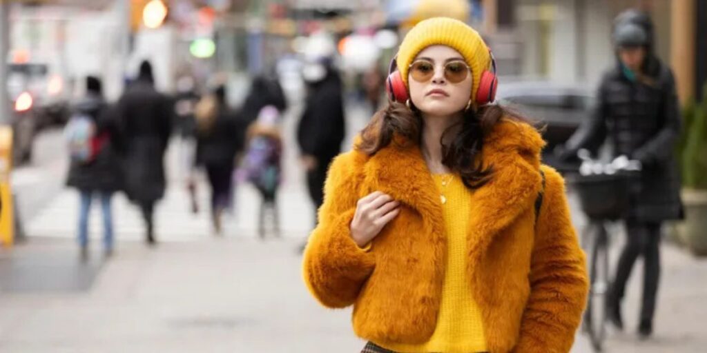 Selena Gomez as Mabel in Only Murders In The Building walking down a road waring sunglasses and a yellow outfit