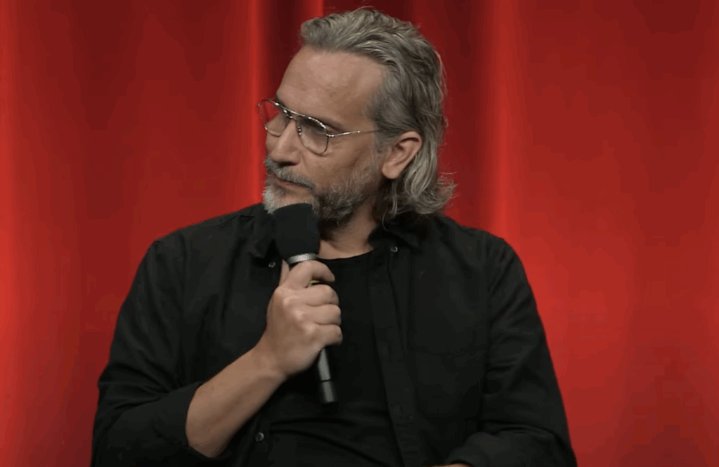 Fede Álvarez holding a mic, wearing a black T, shirt and glasses in an interview.