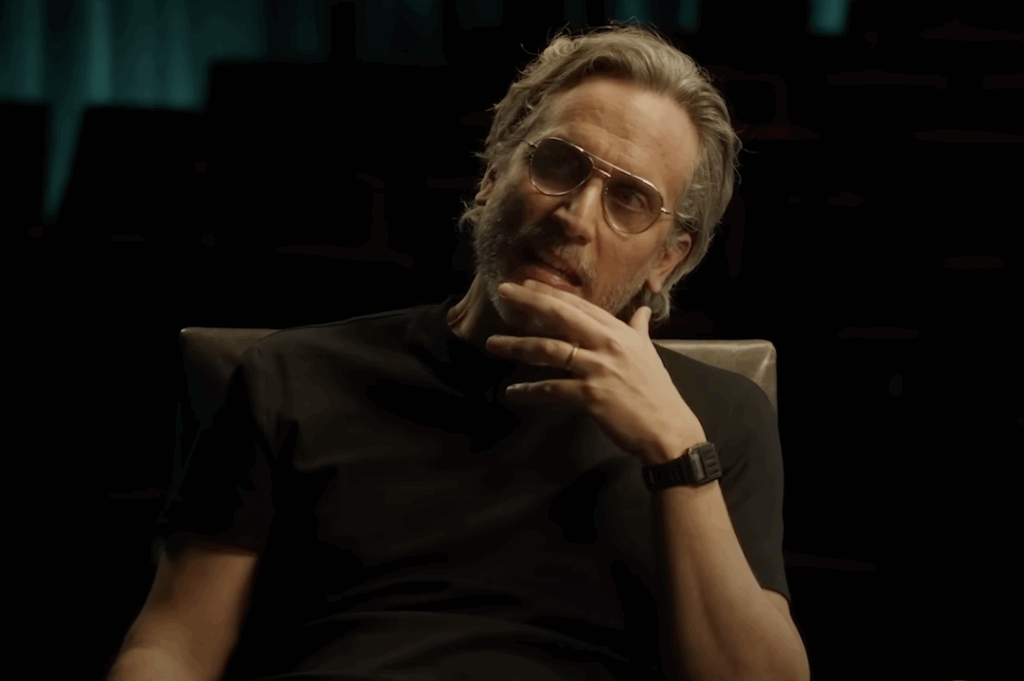 Fede Álvarez wearing a black T, shirt, and glasses, with a hand on his chin during an interview.
