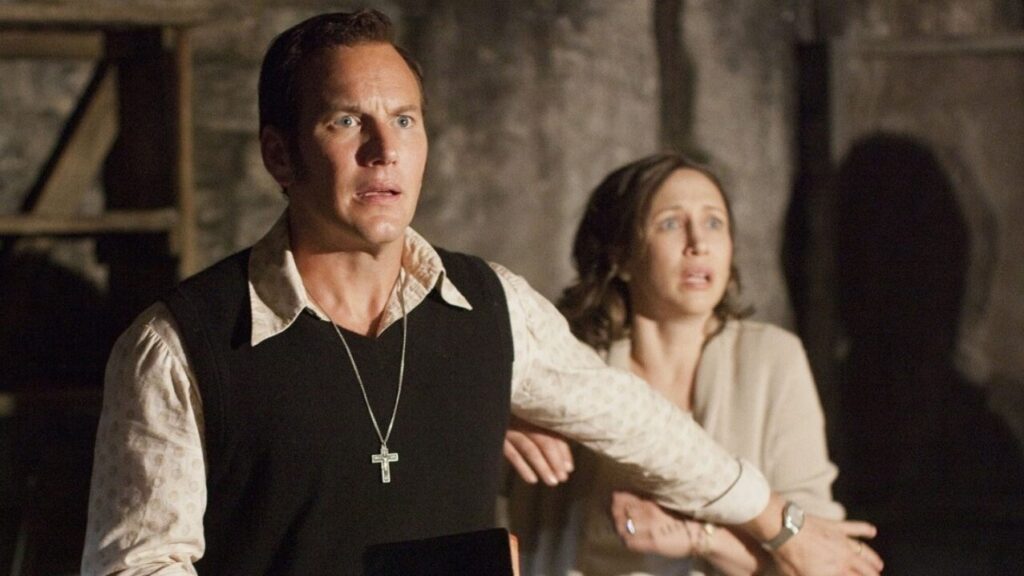 Patrick Wilson as Ed Warren and Vera Farmiga as Lorraine Warren in The Conjuring, both looking concerned while investigating a paranormal case in a dimly lit basement setting in one of the movies from The Conjuring universe.
