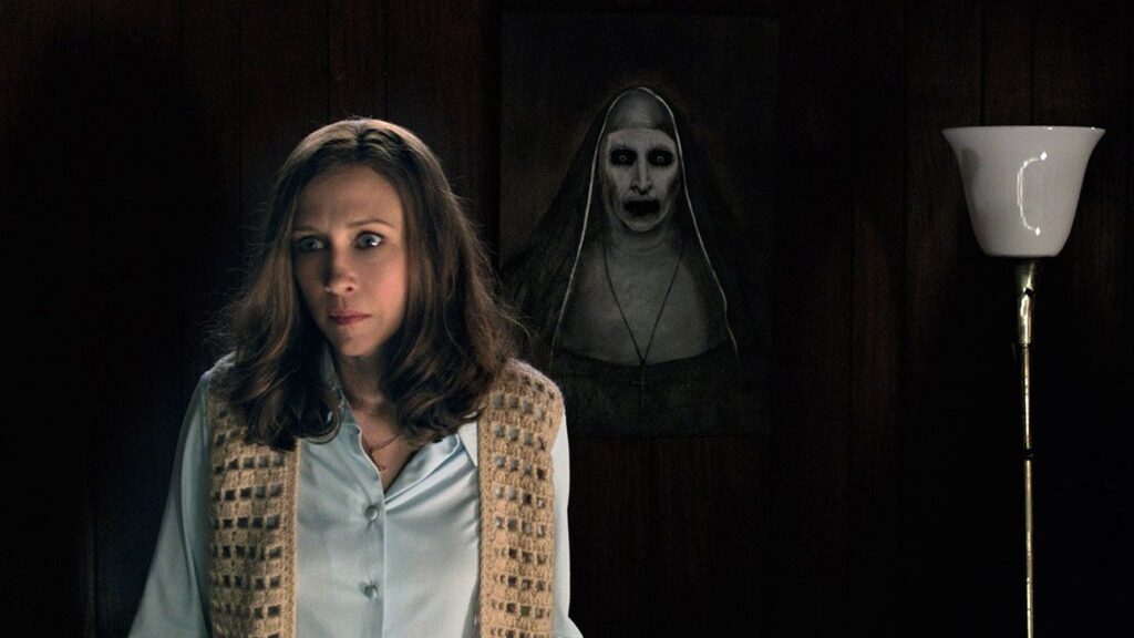 Vera Farmiga as Lorraine Warren in The Conjuring 2 standing in a dark room with the portrait of terrifying demonic Nun (Valak) looming menacingly behind her.