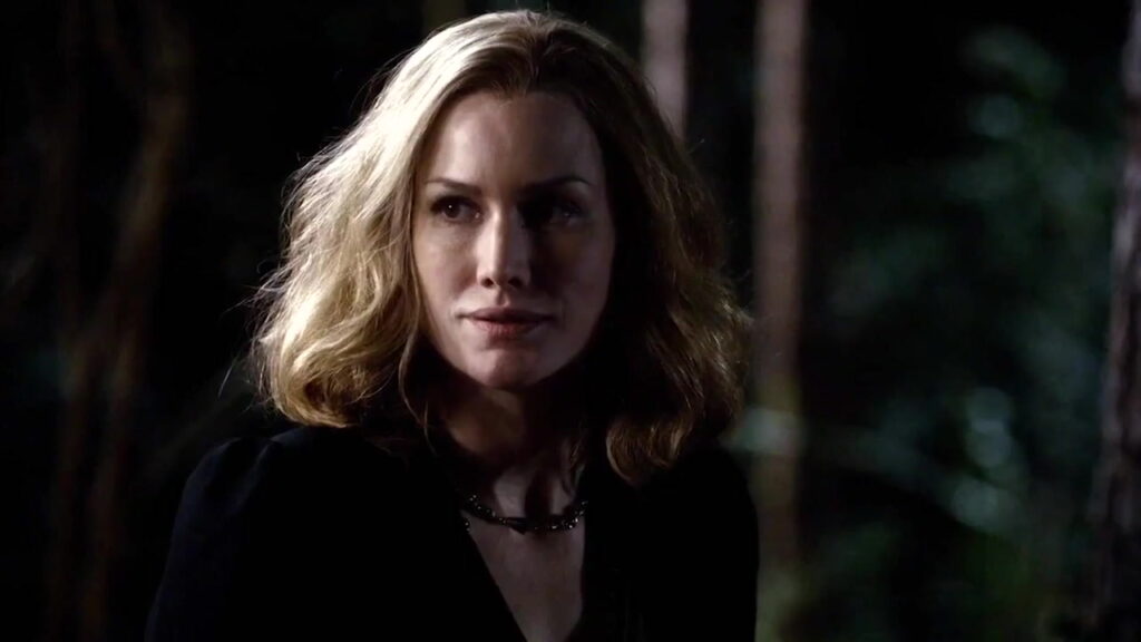 Alice Evans as Esther Mikaelson with shoulder-length blonde wavy hair wearing a black top with a chain necklace, looking to the side with a smirk in dim lighting.