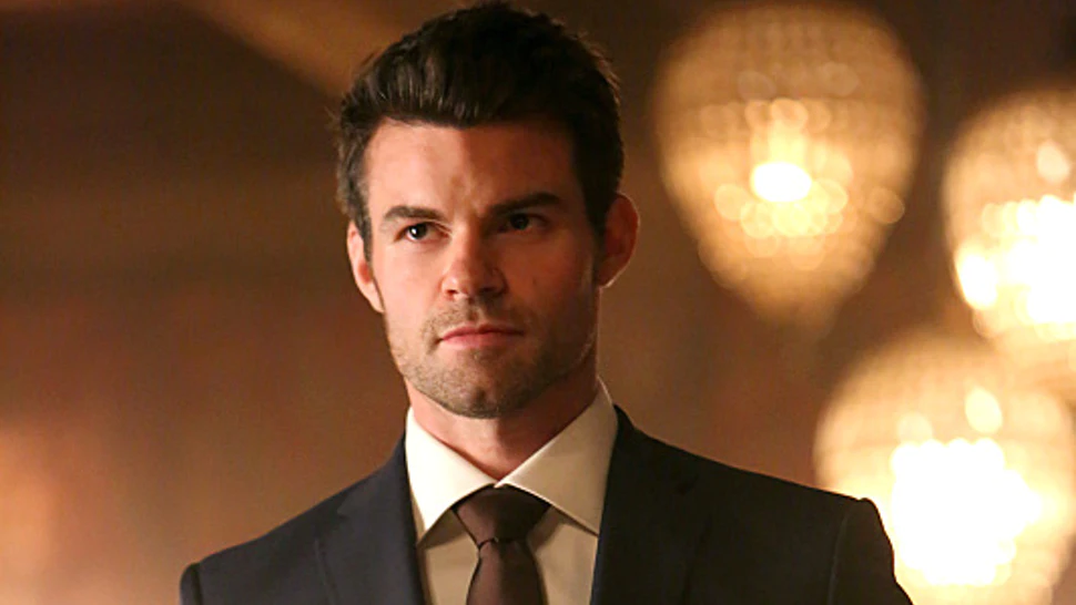 Daniel Gillies as Elijah Mikaelson with dark hair and facial stubble wearing a formal black suit with white shirt and tie, photographed against a background of warm, blurred lights.