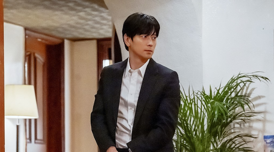 A man in a dark suit and white shirt is standing indoors. He is looking off to the side with a serious expression.