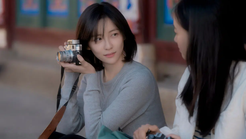 A woman with a short bob haircut is holding a camera and talking to another person. She is looking at the person while holding the camera.