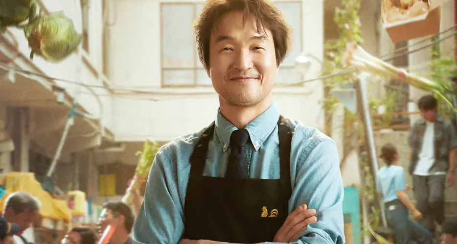 A man with a warm smile is standing in a market. He's wearing an apron over a blue shirt and has his arms crossed.