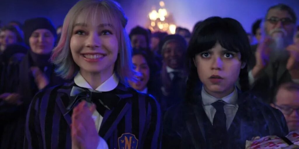 Enid Sinclair smiling and Wednesday Addams looking grim as they stand beside each other in Wednesday