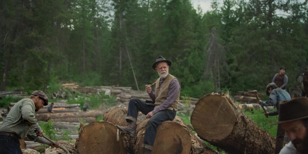 William H. Macy sitting on a log in Train Dreams 