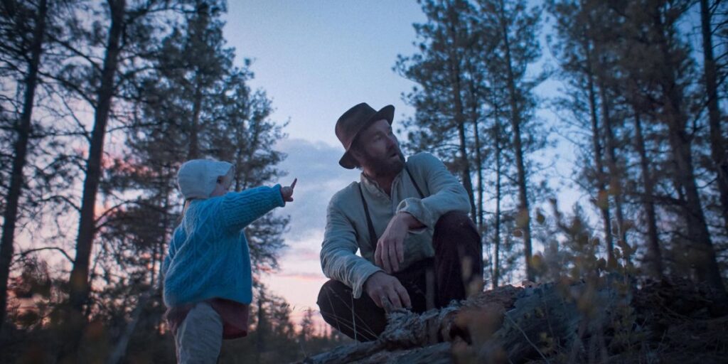 Joel Edgerton crouching in Train Dreams with a baby standing in front of him and pointing at something
