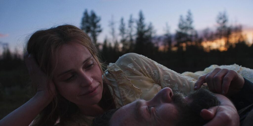 Joel Edgerton lying on grass as Felicity Jones caresses his face in Train Dreams