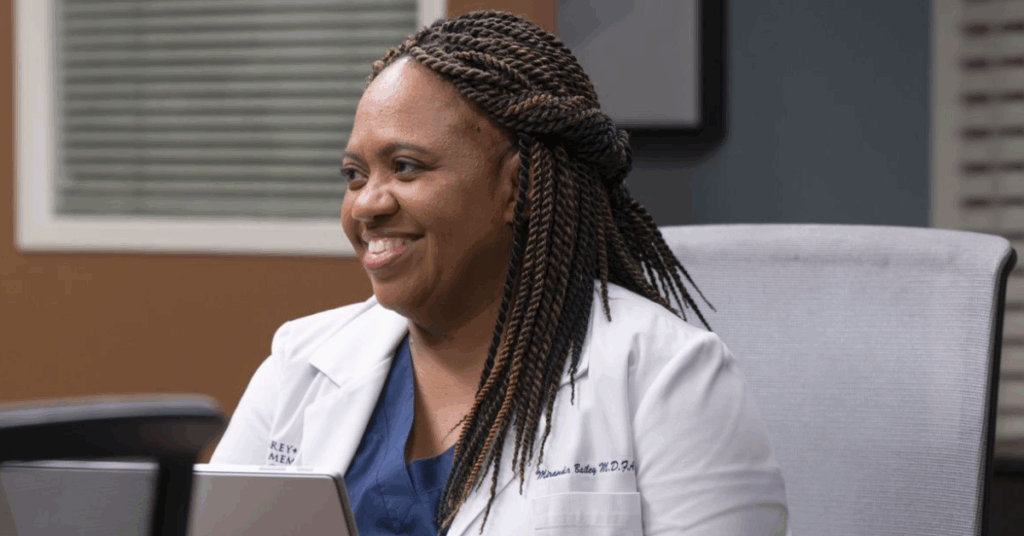 Miranda Bailey sitting in front of a laptop while wearing her doctor's scrub and smiling. 
