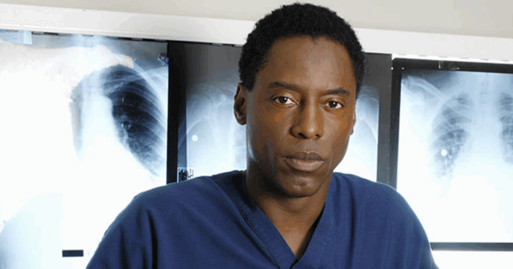 Isaiah Washington looking at something intensely while wearing a blue scrub. 
