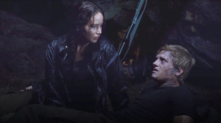In a dark, cavernous setting, a concerned Katniss tends to an injured Peeta who is lying on the ground. 
