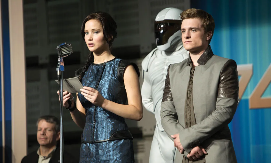 Katniss, holding a paper, stands at a microphone next to Peeta, with a Capitol guard behind them.
