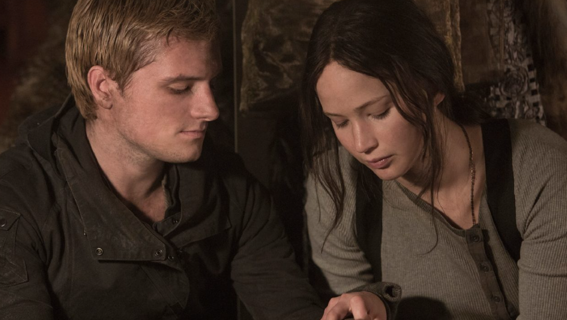 Peeta and Katniss are sitting closely together, looking down at something in front of them with serious expressions. 