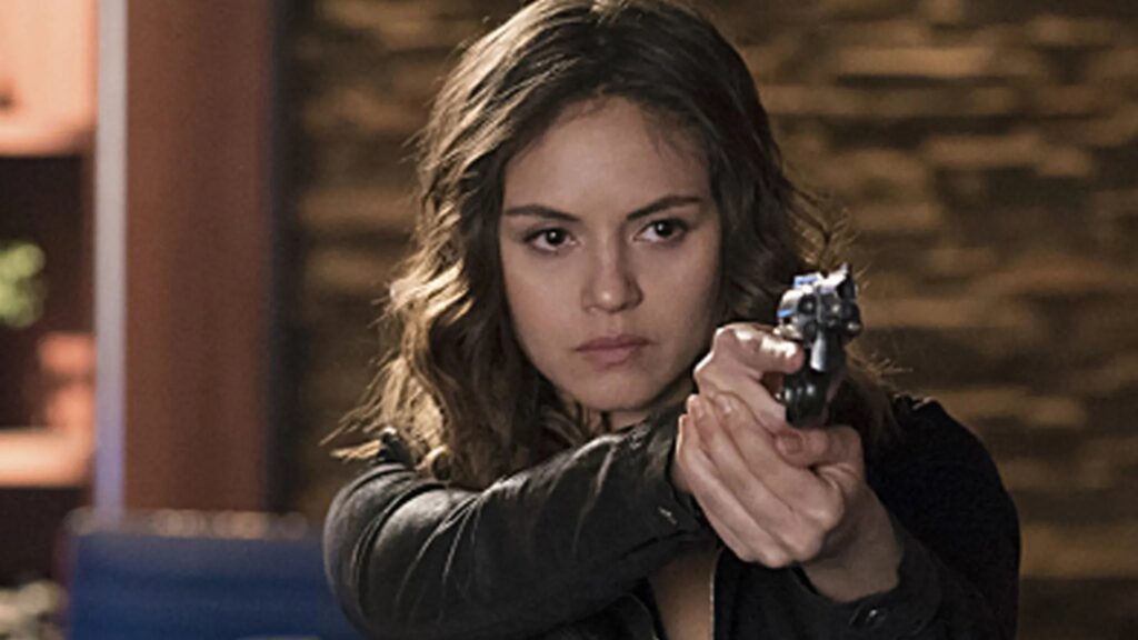 Leslie-Anne Huff as Rayna Cruz with wavy brown hair wearing dark clothing, pointing a gun directly at the camera with a serious, determined expression.