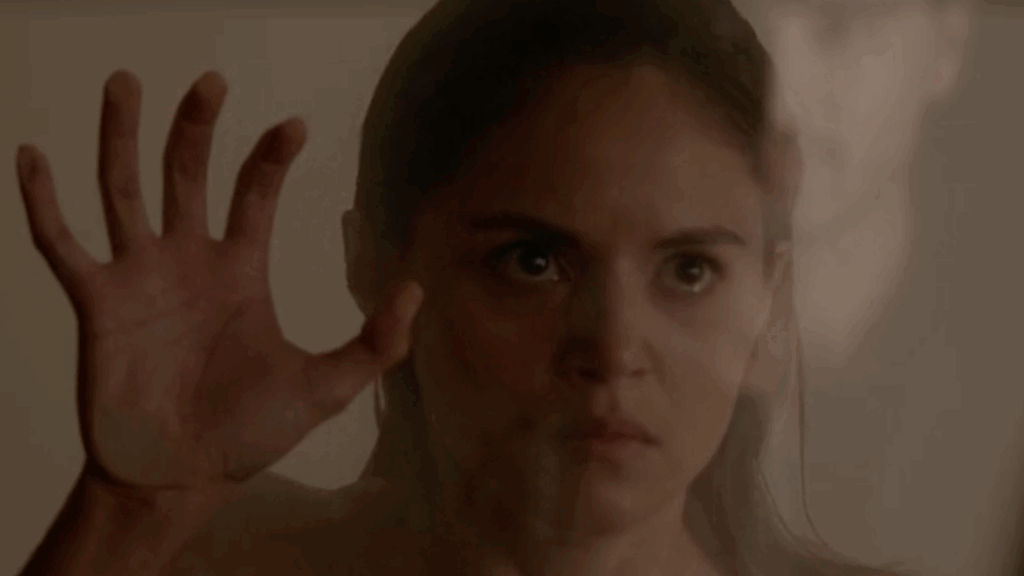 Leslie-Anne Huff as Rayna Cruz raising her hand toward the camera with her palm placed on the glass wall behind which she is standing, with an intense expression on her face.
