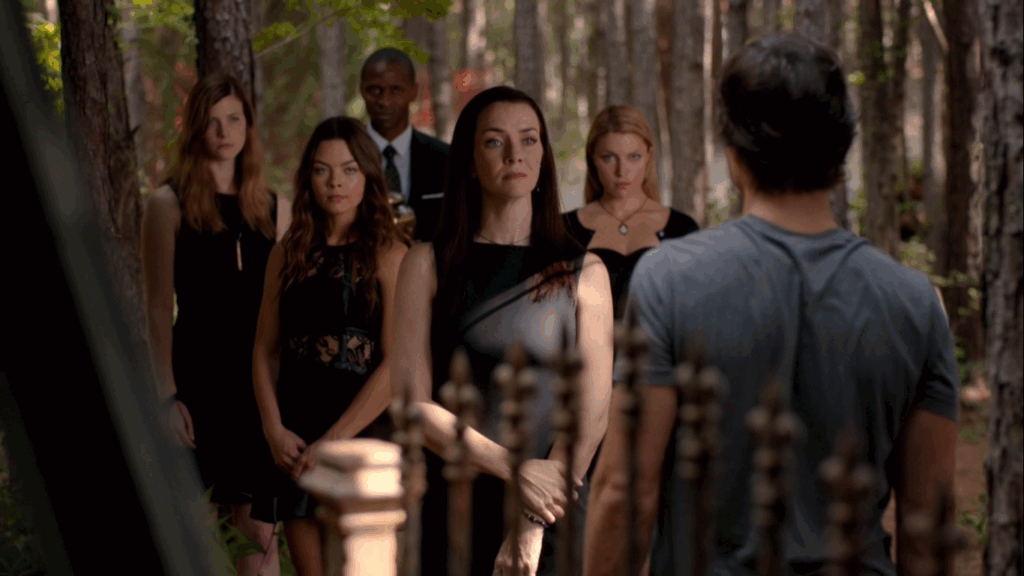 The Heretics dressed in black standing together outdoors among trees, with Damon Salvatore in the foreground facing away from the camera toward the Heretics.