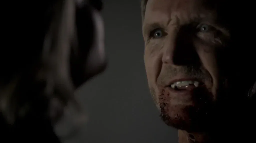 Sebastian Roché as Mikael with light eyes and facial hair looking at someone facing away from the camera with an intense, frightening expression, with dark veins visible around his eyes and dried blood on his chin, photographed in dramatic lighting.