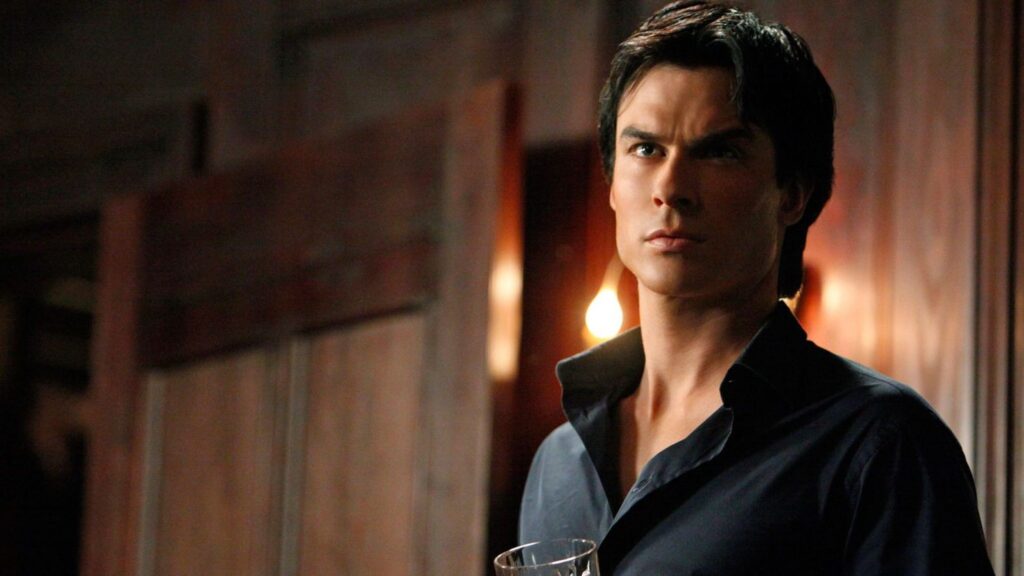 Ian Somerhalder as Damon Salvatore with dark hair wearing a dark shirt, holding a glass and looking serious in a dimly lit interior with candles visible in the background.