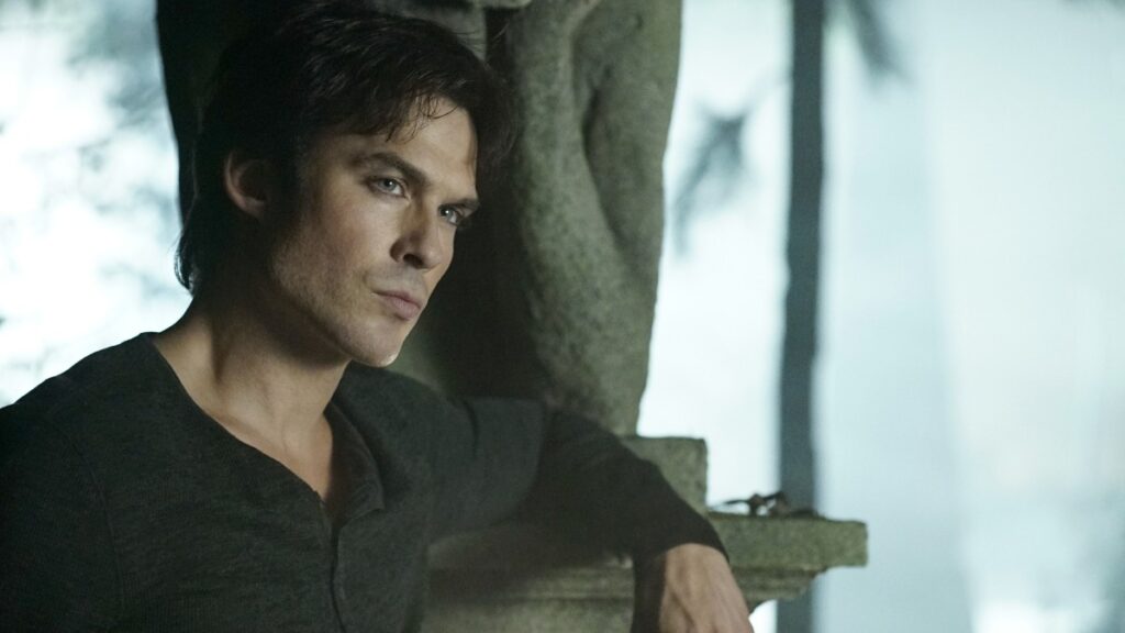 Ian Somerhalder as Damon Salvatore with dark tousled hair wearing a dark henley shirt, leaning against a stone pillar in a crypt or stone building.