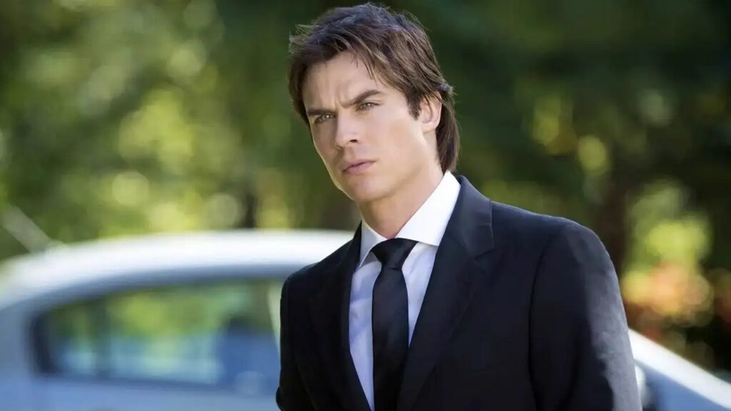 Ian Somerhalder as Damon Salvatore with dark hair wearing a formal black suit with white shirt and dark tie, standing outdoors with a car and trees visible in the blurred background.