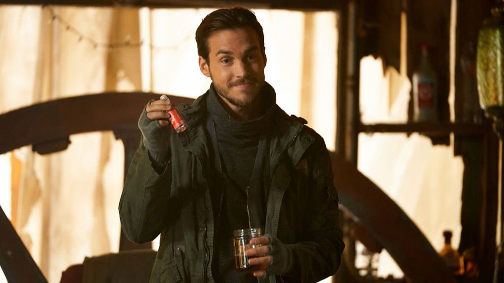 Chris Wood as Kai Parker with dark hair wearing a dark green jacket, smiling while holding a red bottle and glass in a rustic indoor setting with warm lighting.