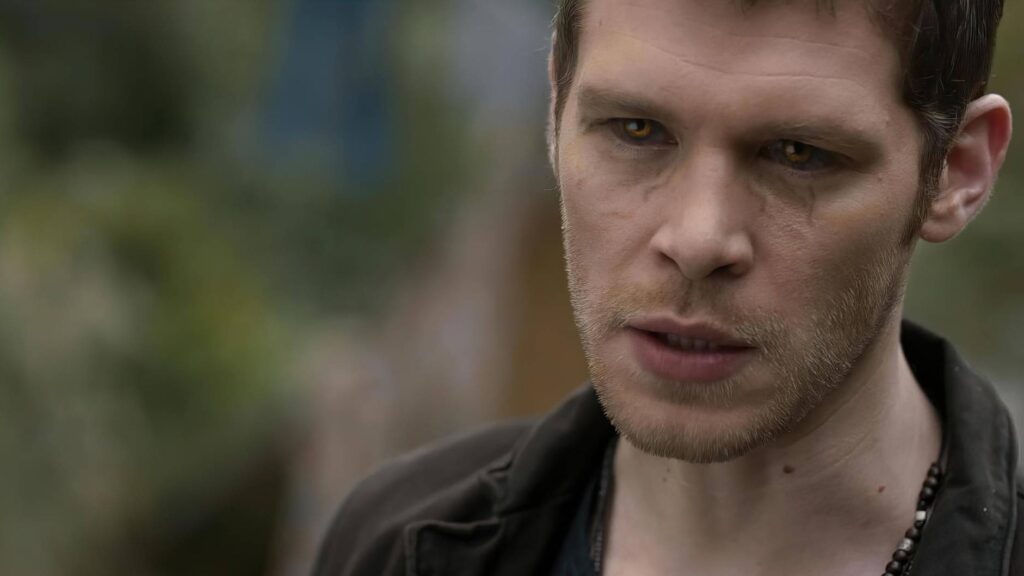 Joseph Morgan as Klaus Mikaelson with short dark hair and stubble wearing a dark shirt, photographed outdoors with a serious, intense expression.