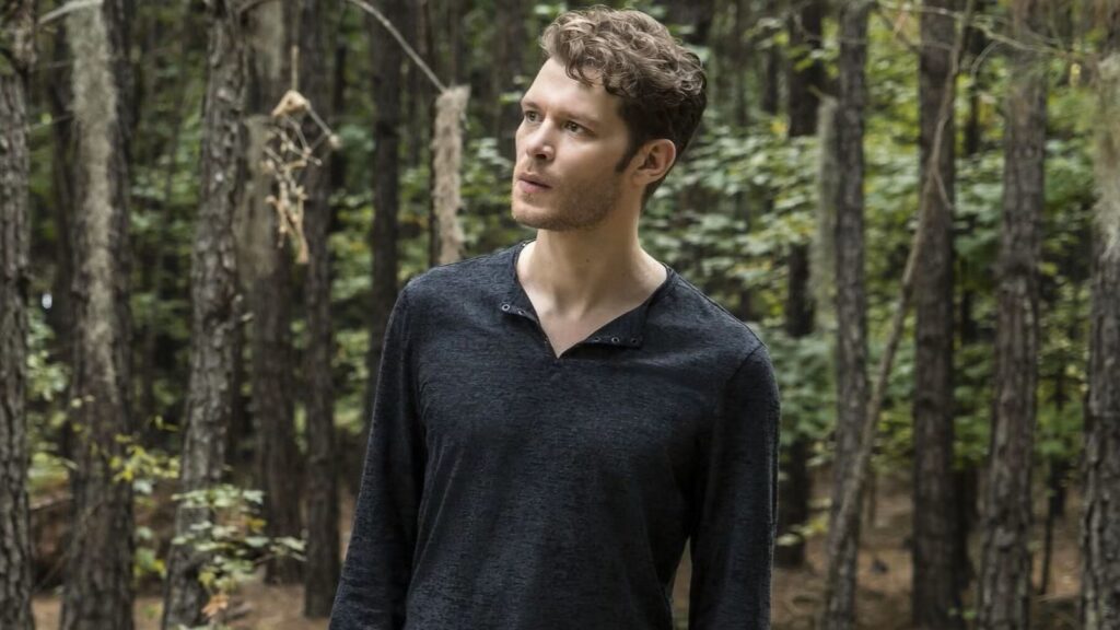 Joseph Morgan as Klaus Mikaelson with wavy dark hair wearing a dark long-sleeved shirt, standing outdoors in a wooded area during daylight.