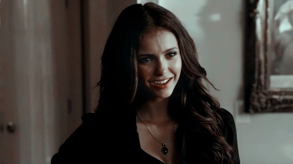 Nina Dobrev as Katherine Pierce with long dark wavy hair wearing a black top and necklace, smiling confidently in an indoor setting.