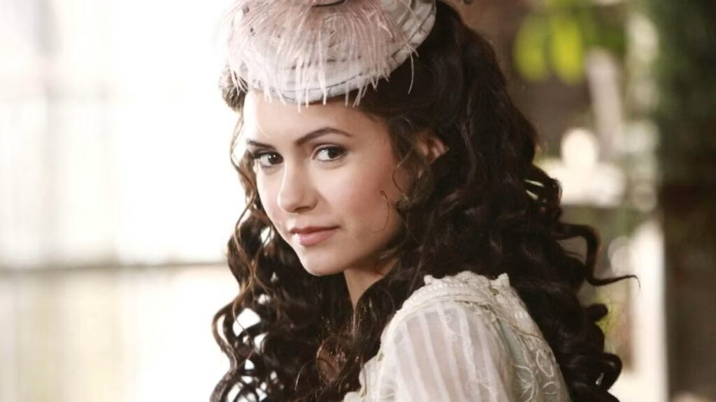 Nina Dobrev as Katherine Pierce with dark hair wearing a white period dress with puffy sleeves and a white feathered hat, photographed in profile.